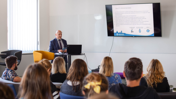 People sit with their backs to the lens on chairs and watch the presentation projected in front of them , which is described by a man in a suit who stands on the left side of the photo